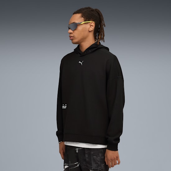 A model in a black hoodie and sunglasses stands against a grey backdrop.