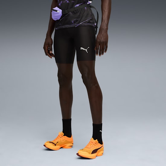 Against grey, a model wears a patterned top, black shorts, and orange running shoes.