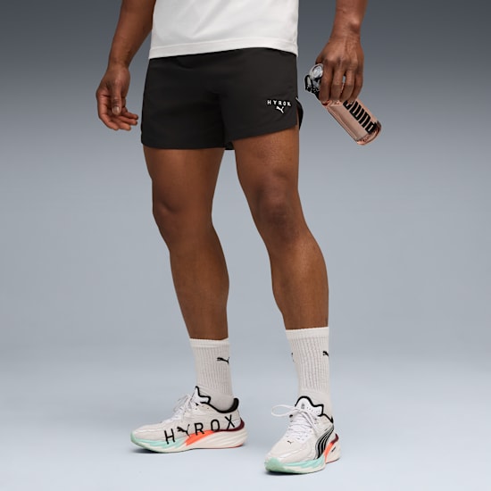 A model on grey, wearing a white t-shirt, black shorts, running shoes and holding a bottle.