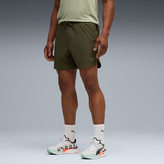 A model poses against a grey backdrop, wearing a green top, shorts and running shoes.