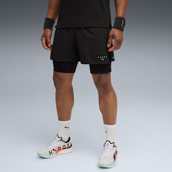 A model is standing against a grey background wearing black sportswear and running shoes.