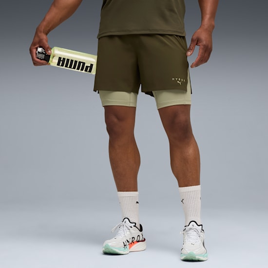Against grey, a model holds a bottle, wearing an olive top, shorts, white socks and running shoes.