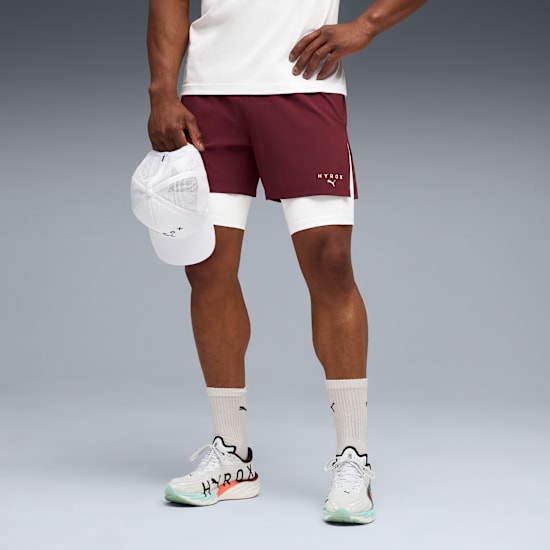 A model poses against grey wearing shorts, shoes and holding a white cap.