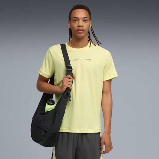 On a grey background, a model wears a yellow tee, grey shorts and a black bag.