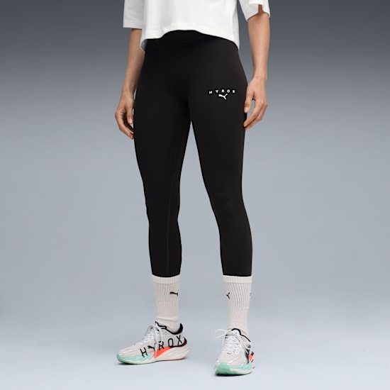 A model poses in front of grey wearing black leggings, trainers, and a white t-shirt.