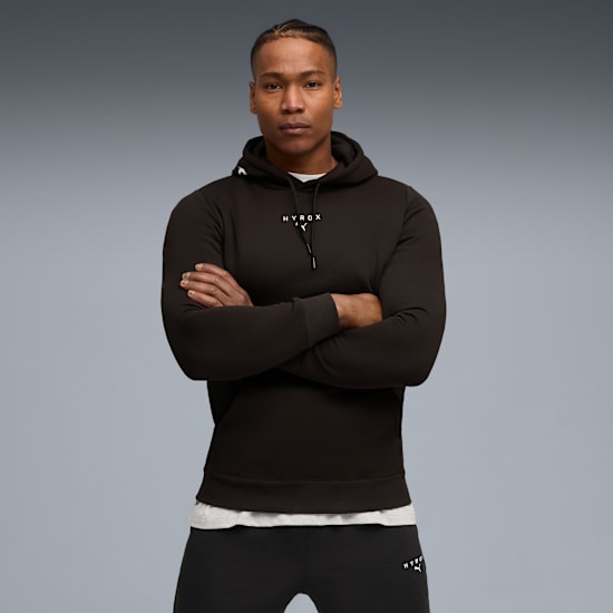 Tegen een grijze achtergrond poseert een model met gekruiste armen in zwarte hoodie en broek.