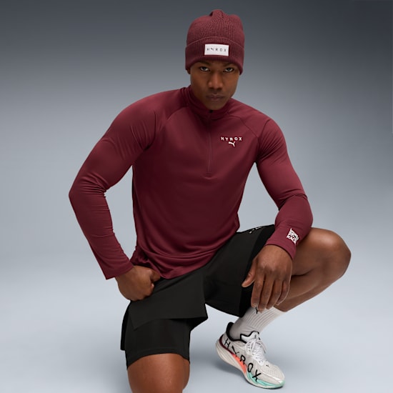 A model in front of grey background is crouching and wearing sportswear with beanie.