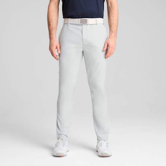 A model poses against a grey backdrop, wearing light grey trousers, white belt and white trainers.