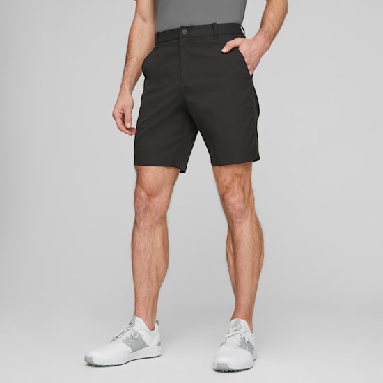 Vor grauem Hintergrund posiert ein Model mit der Hand in der Tasche, mit dunklen Shorts und weißen Turnschuhen.