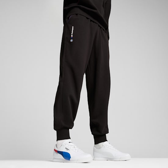 Against a grey backdrop, a model wears black joggers and white trainers.