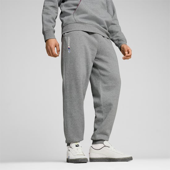 Un modèle sur fond gris porte un pantalon de survêtement, un sweat à capuche gris et des baskets blanches.