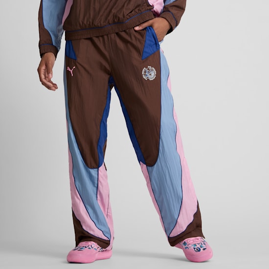 On grey, a model poses in brown, blue, and pink trousers with a hand in pocket.