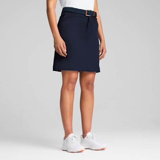 Against a grey backdrop, a model wears a navy skirt with belt and white trainers.