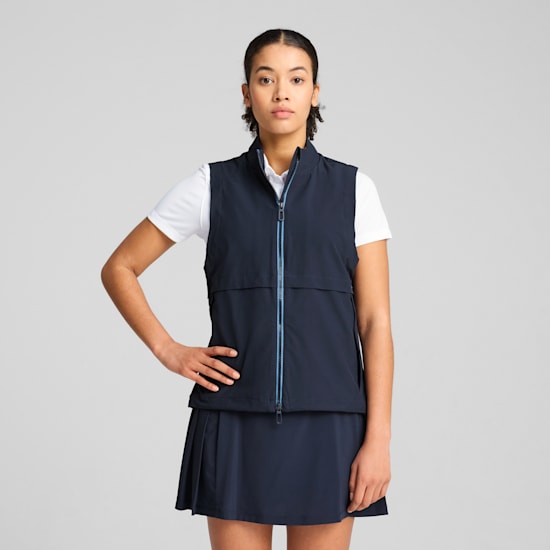 A model poses in front of a grey background wearing a navy vest, skirt and white top.