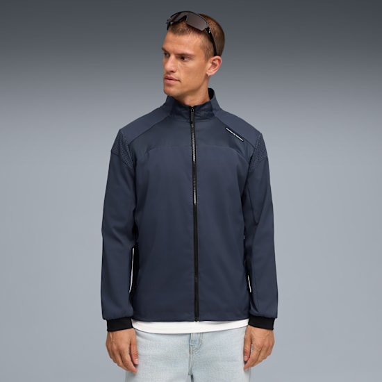 Ein Model vor grauem Hintergrund, trägt eine marineblaue Jacke, helle Jeans und Sonnenbrille.