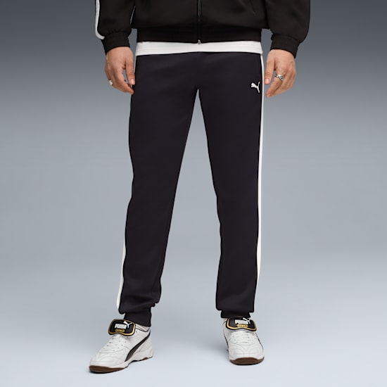 A model stands against grey wearing black tracksuit bottoms with a white stripe and white trainers.