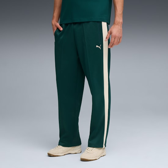 Against grey, a model wears green trousers with a cream stripe and matching trainers.