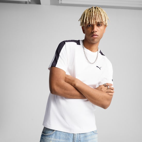 A model with crossed arms poses in front of a grey background wearing a white t-shirt.