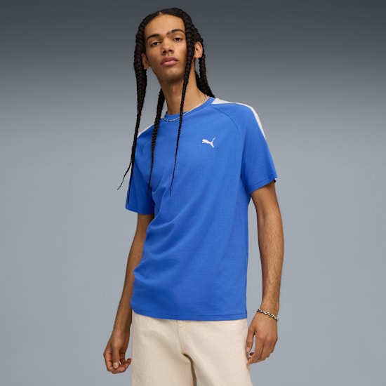 Un modello con trecce posa su sfondo grigio con t-shirt blu e pantaloncini color crema.