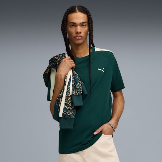 Vor grauem Hintergrund posiert ein Model mit grünem T-Shirt, heller Hose und Jacke über der Schulter.