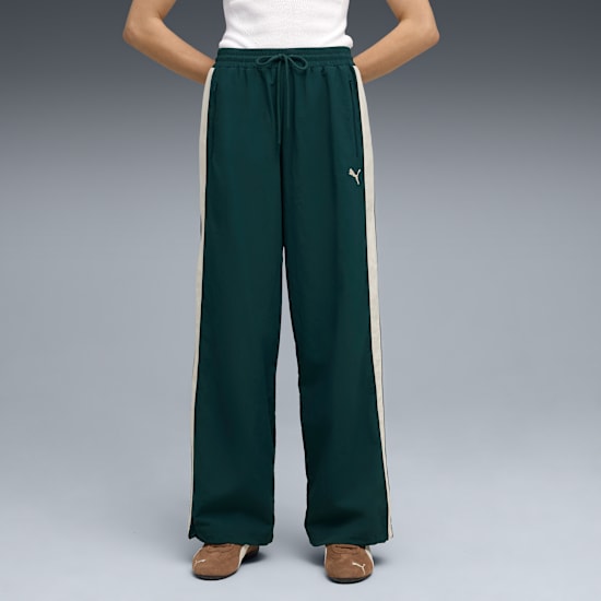 Against grey, a model poses in green trousers with side stripes and brown trainers.