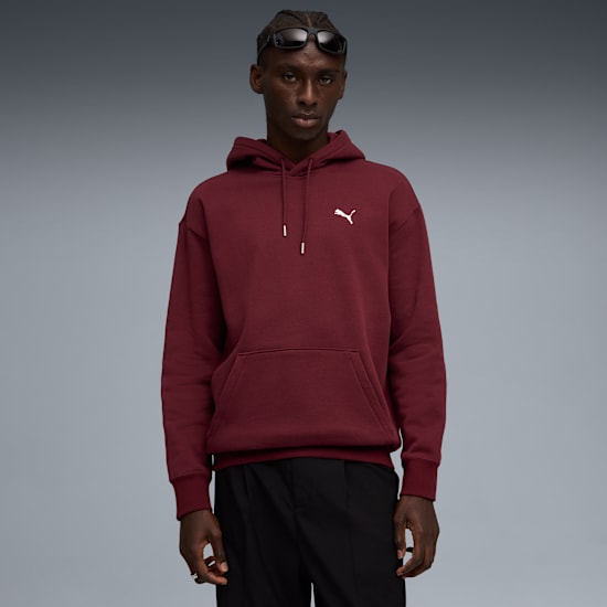 Against grey, a model poses in a burgundy hoodie and black trousers, with sunglasses.
