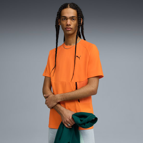 A model with braids poses against grey, wearing an orange t-shirt and holding a green top.