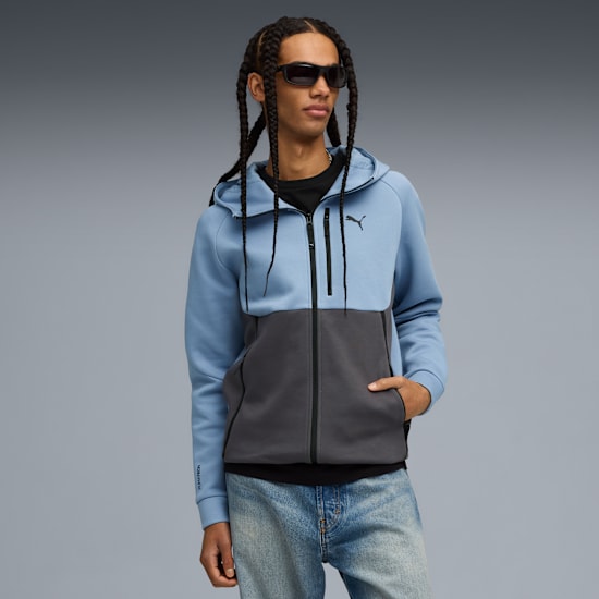 A model with braids wears a two-tone zip-up hoodie and sunglasses against a grey background.