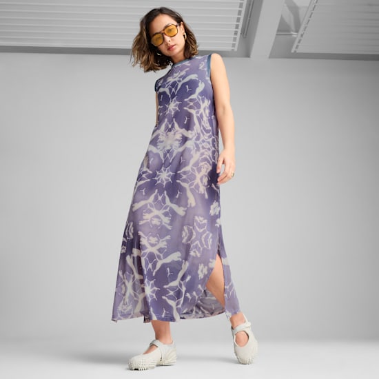 A model poses on grey wearing a patterned dress, light shoes, and yellow tinted glasses.