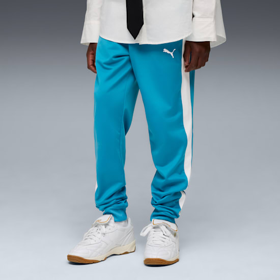 Against grey, a model wears teal trousers, white trainers, and a white shirt with a black tie.