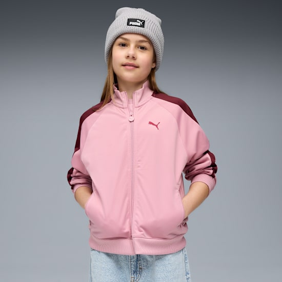 Ein Model posiert vor grauem Hintergrund mit Mütze, rosa Jacke mit Burgunder Akzenten und Jeans.
