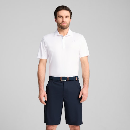 Een model staat tegen een grijze achtergrond, gekleed in een witte polo en donkere shorts.