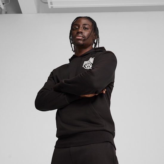 A model with arms crossed in front of a grey backdrop wearing a black hooded top.