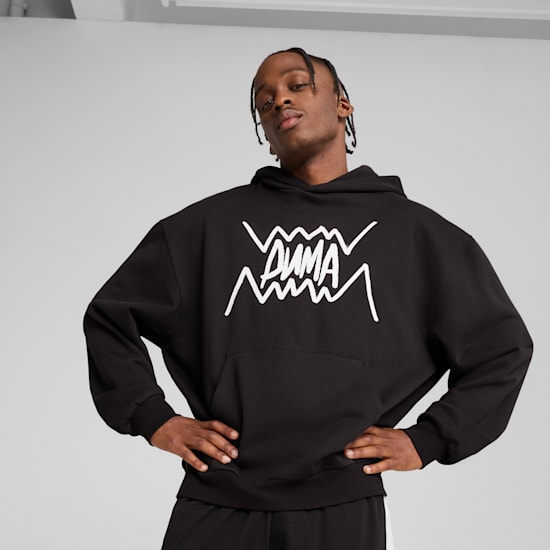 Against a grey backdrop, a model poses in a black hoodie and black bottoms.