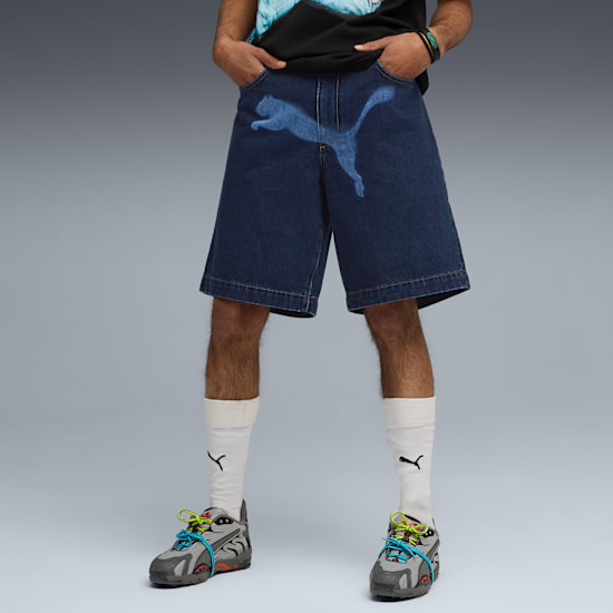 Vor grauem Hintergrund posiert ein Model in Logo-Jeansshorts, Socken und Turnschuhen.