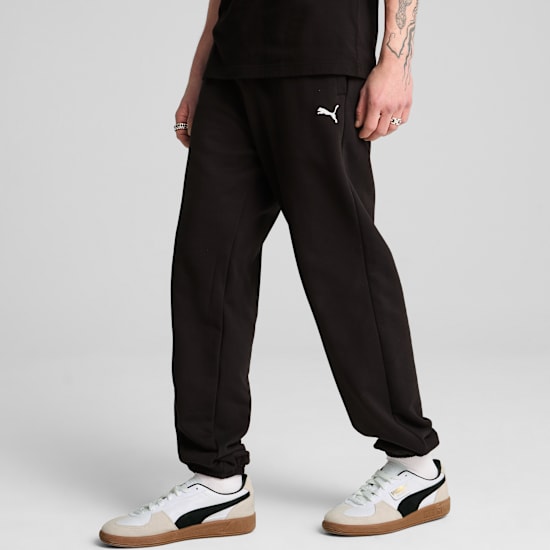 A model poses against a grey backdrop, wearing black trousers and white and black trainers.