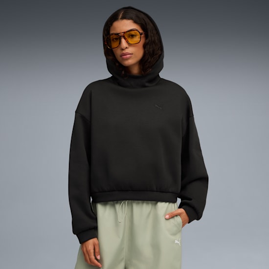 Un modello con occhiali posa su sfondo grigio, indossando felpa nera con cappuccio e pantaloni verdi.