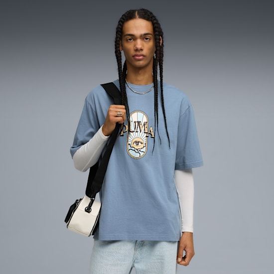 Op grijs draagt een model een blauw T-shirt met print, lichte jeans en een tas.