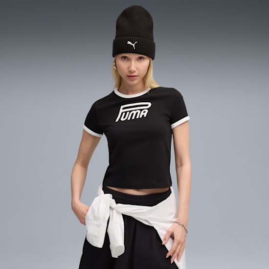 A model in front of a grey background wearing a black t-shirt, beanie and black skirt.