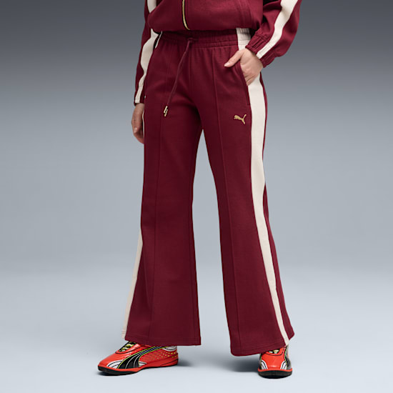 Against grey, a model poses wearing maroon trousers with cream stripes and red trainers.