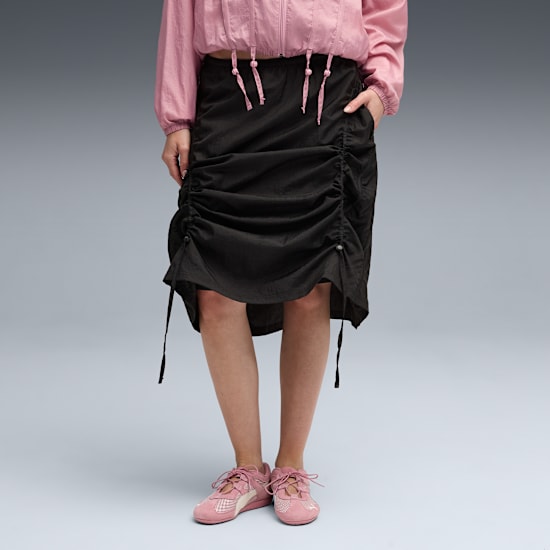 Against grey, a model wears a pink jacket, black skirt and pink trainers.