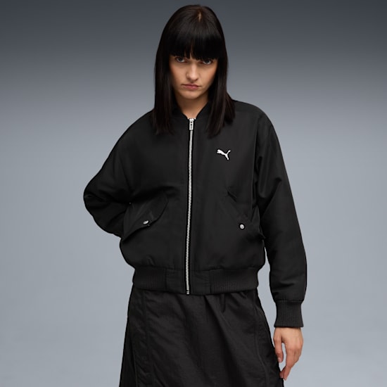 Een model poseert voor grijs, gekleed in een zwart jack met wit logo en een zwarte rok.