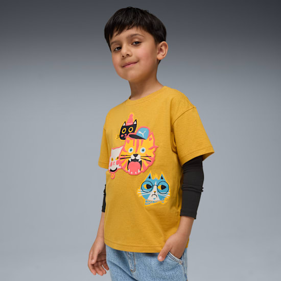 A child poses against grey backdrop, wearing a yellow tee with cat designs and blue jeans.