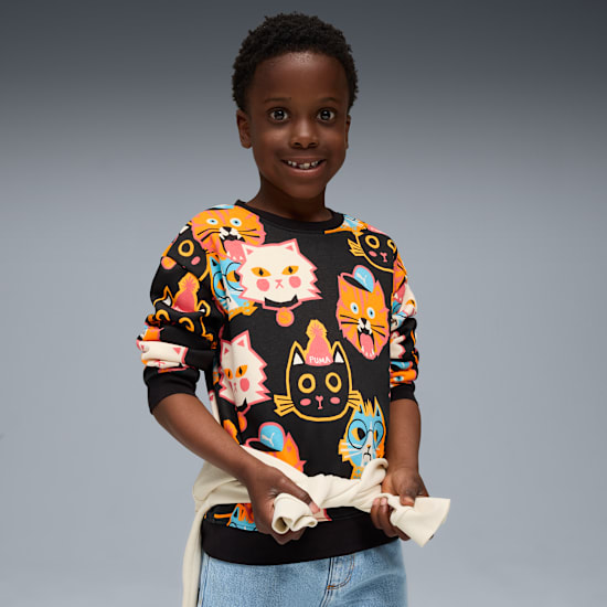 Sur fond gris, un enfant souriant en sweat noir à imprimé chat tient un haut crème.