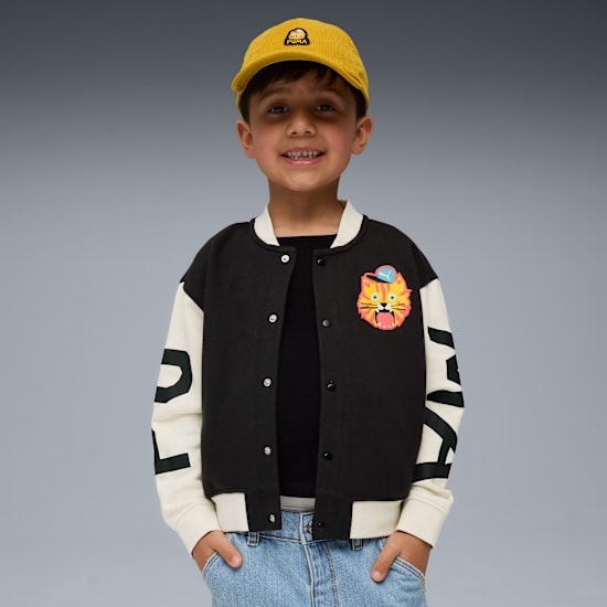 Ein Kind posiert vor grauem Hintergrund mit gelber Kappe, Jacke, schwarzem Oberteil und Jeans.