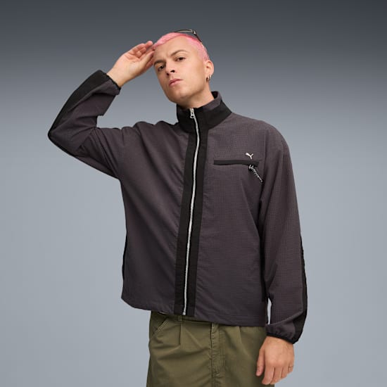 Vor grauem Hintergrund posiert ein Model mit pinken Haaren in grauer Jacke und grüner Hose.