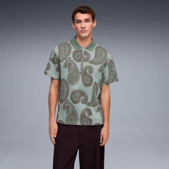 A model stands against a grey backdrop, wearing a paisley patterned polo shirt and dark trousers.