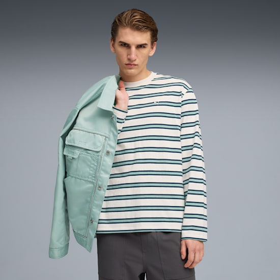 A model poses against grey, wearing a striped top, grey trousers and holding a light green jacket.