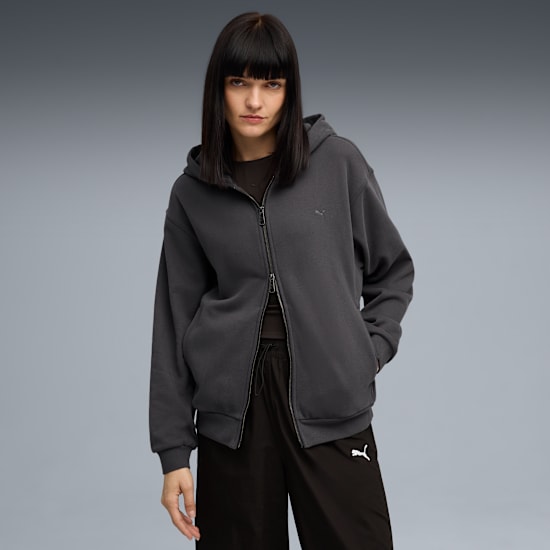 A model poses against a grey background, wearing a zipped hoodie and black trousers.