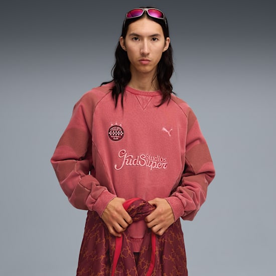 A model stands against a grey backdrop, wearing a pink sweater and patterned trousers.